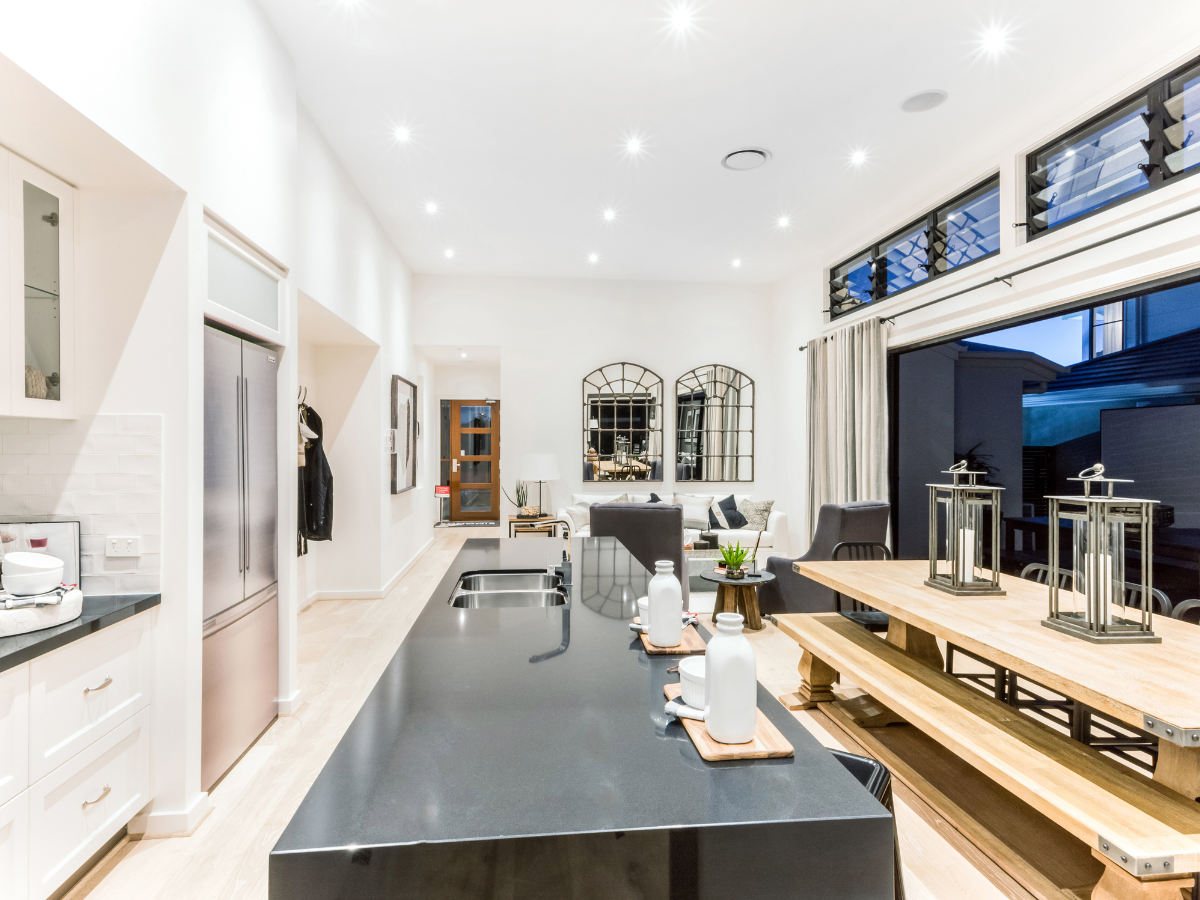 Modern open-plan integrated kitchen and living area with black island, wooden dining table, bench seating, and large windows. Neutral decor and recessed lighting create a bright, spacious atmosphere.