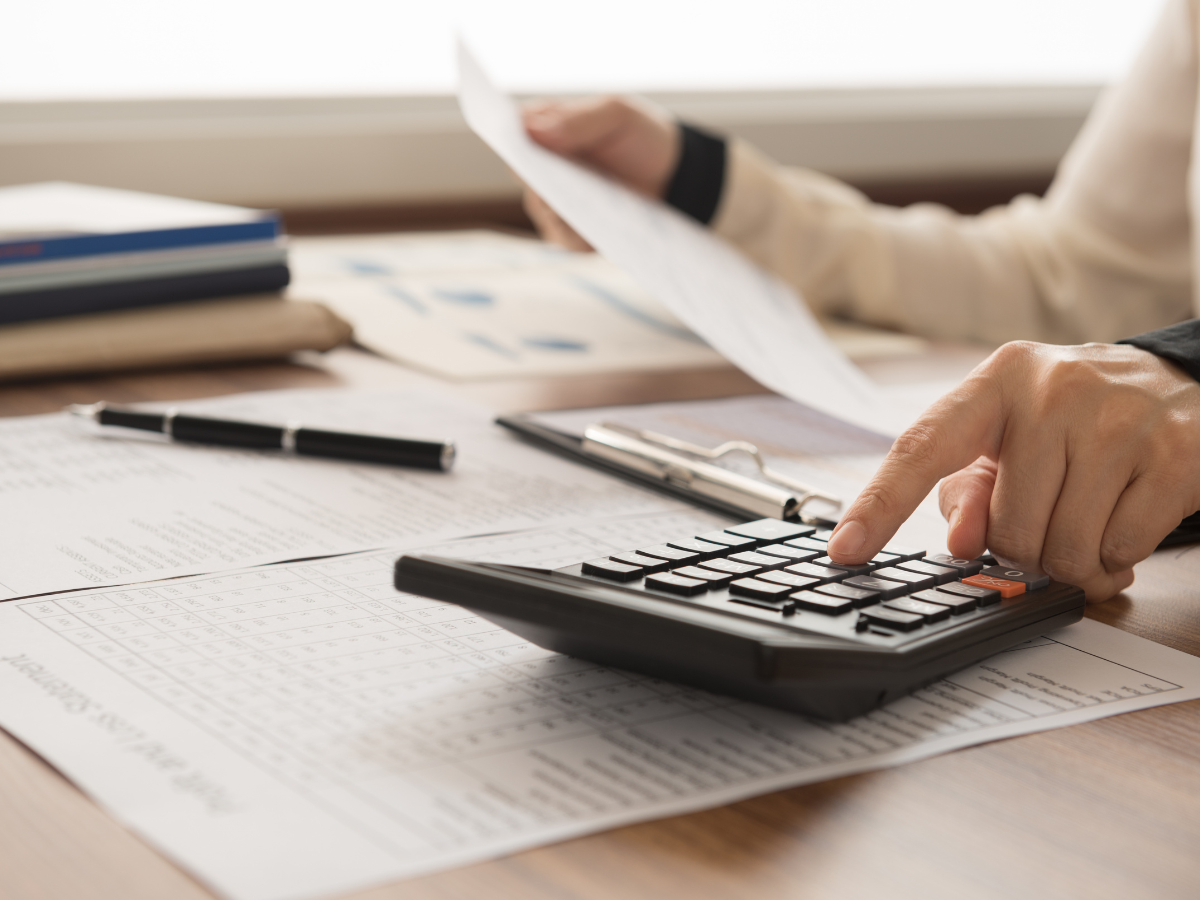 DIY Kitchen Remodeling Tips for a Successful Makeover 3 Person using a calculator with financial documents, pen, and clipboard on a wooden desk while planning DIY kitchen remodeling expenses. One hand holds a sheet of paper.