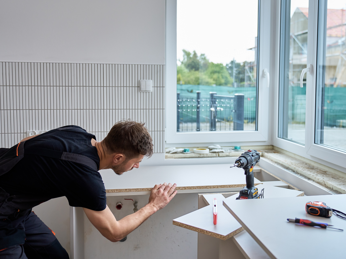 DIY Kitchen Remodeling Tips for a Successful Makeover 5 A man in work overalls installs a countertop in a kitchen, with tools such as a drill and a level on a nearby surface, showcasing the hands-on approach of DIY kitchen remodeling.