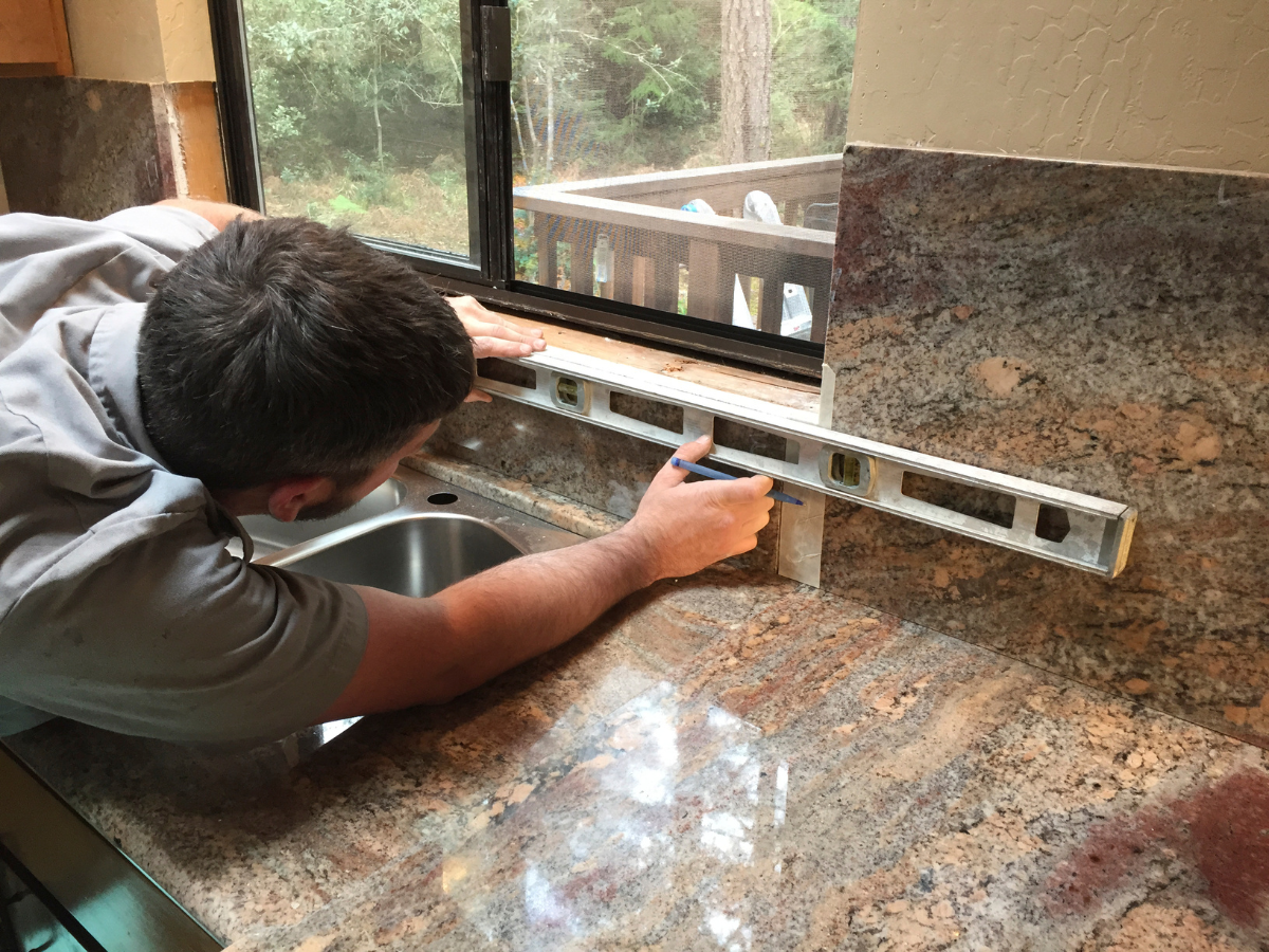 DIY Kitchen Remodeling Tips for a Successful Makeover 6 A person uses a spirit level to check alignment while installing a backsplash above a kitchen sink, next to a window—a perfect example of hands-on DIY kitchen remodeling.