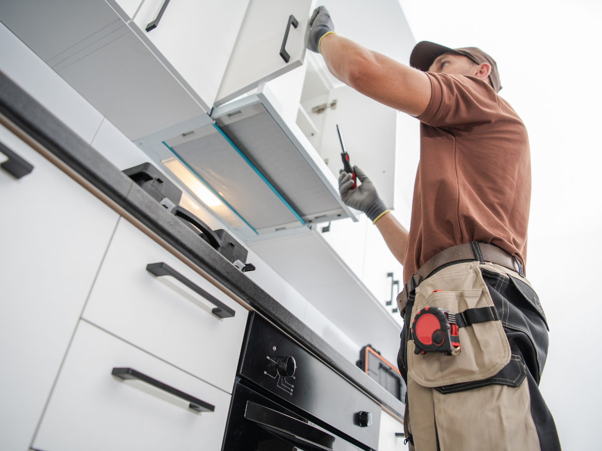 DIY Kitchen Remodeling Tips for a Successful Makeover 8 A worker in a brown shirt and tool belt uses a screwdriver to install or fix a white kitchen cabinet above a stove, showcasing the hands-on approach of DIY kitchen remodeling.