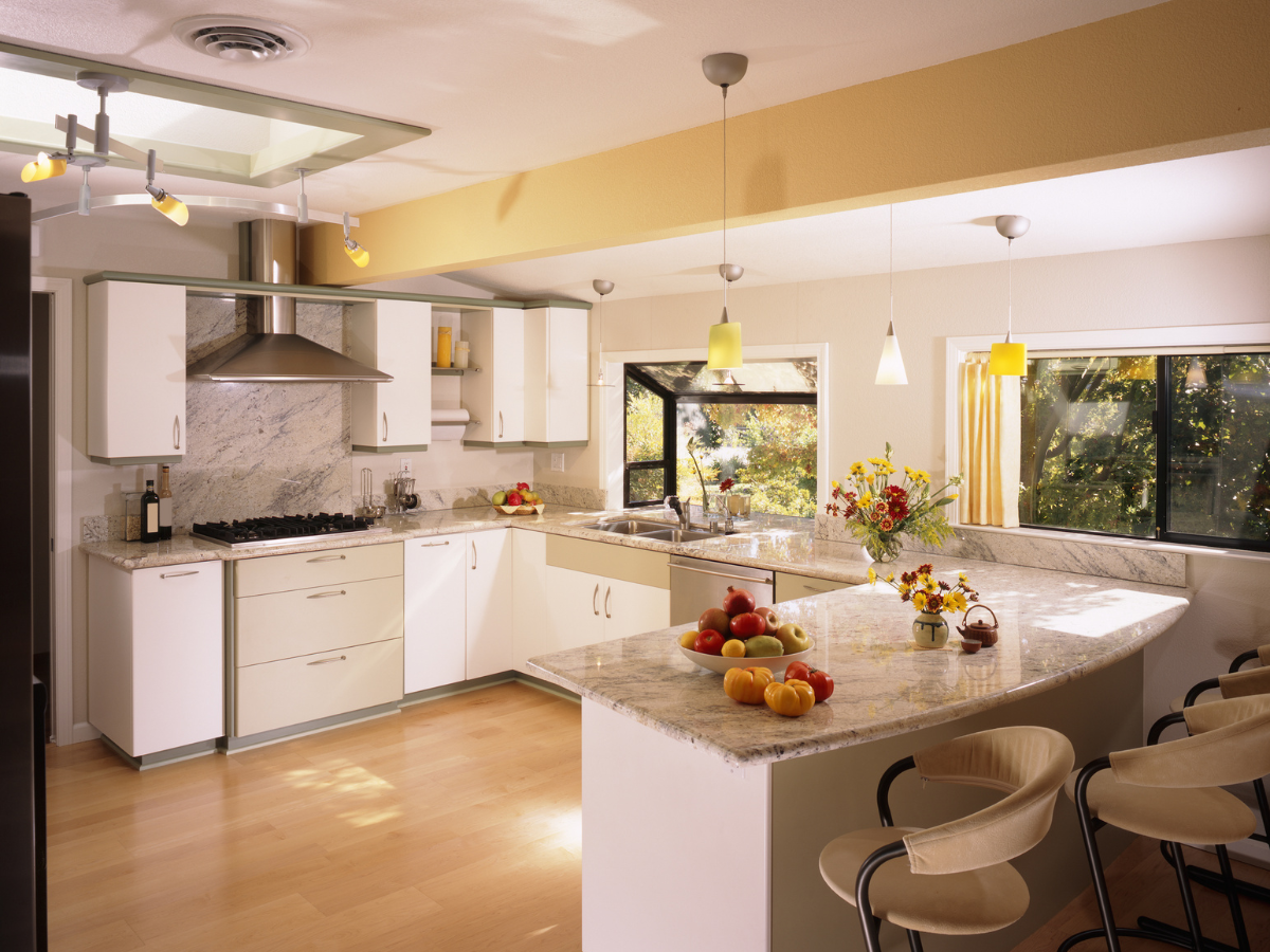 DIY Kitchen Remodeling Tips for a Successful Makeover 1 Modern kitchen with white cabinets, marble countertops, barstools, stainless steel appliances, pendant lights, and bowls of fresh fruit on the counter. Large windows provide natural light—perfect inspiration for your next DIY kitchen remodeling project.