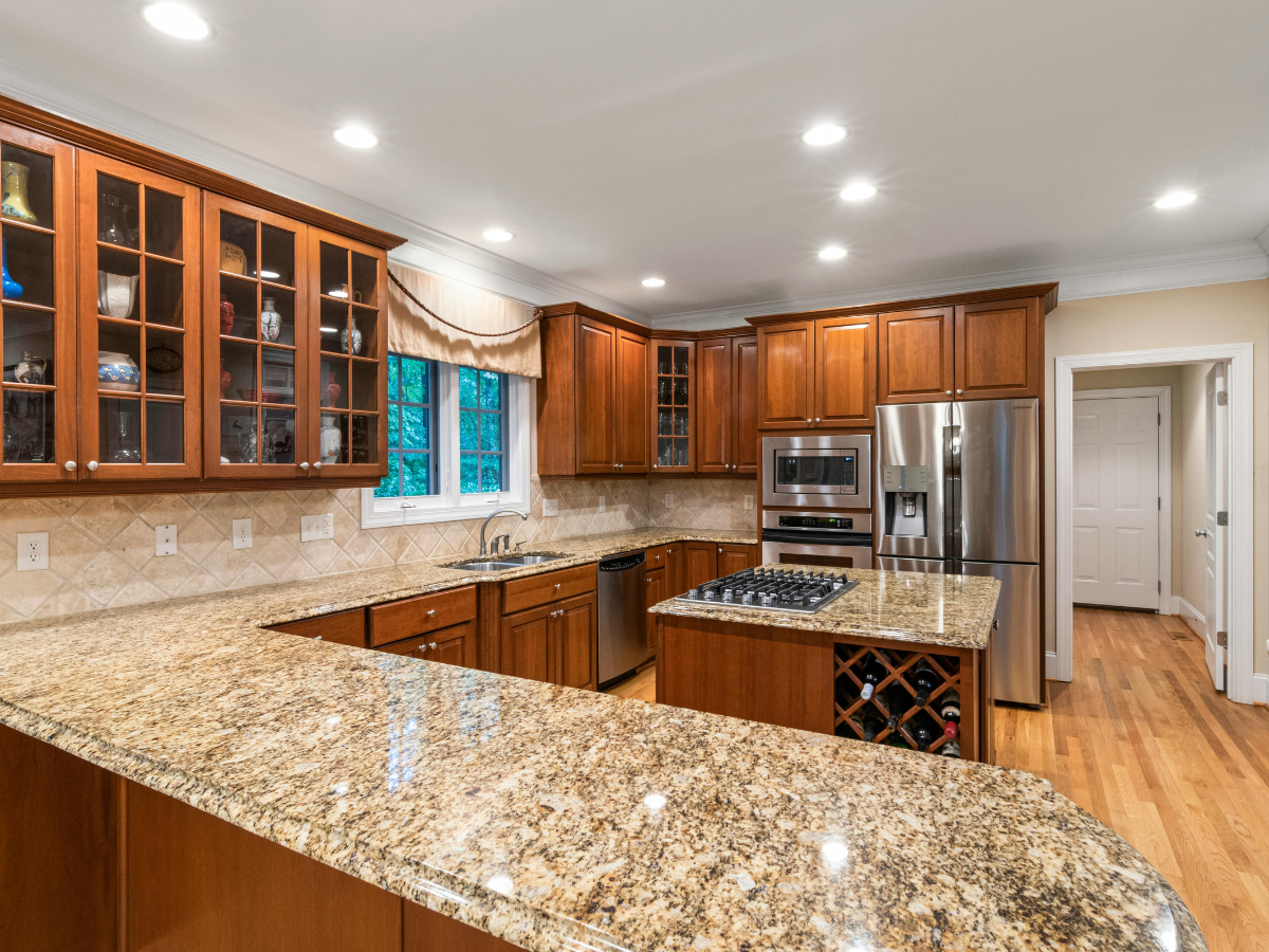 A Countertop Seams Guide to Fabrication and Installation 1 Spacious kitchen with wooden cabinets, granite countertops featuring expertly crafted countertop seams guide for a flawless finish, built-in stainless steel appliances, island with wine rack, and hardwood flooring under recessed lighting.