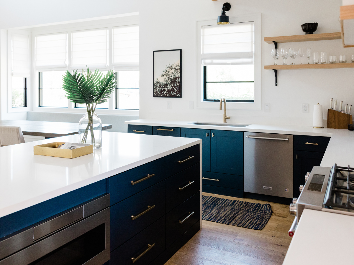 A Countertop Seams Guide to Fabrication and Installation 2 Modern kitchen with navy cabinets, white countertops featuring seamless finishes inspired by the latest countertop seams guide, stainless steel appliances, a vase with palm leaves, and open shelves with glassware. Bright natural light from windows.