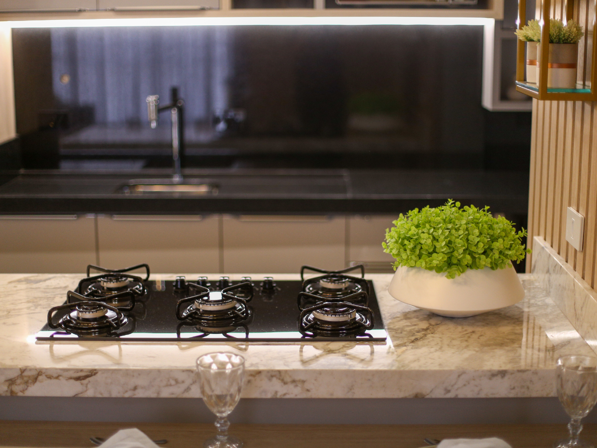 A Countertop Seams Guide to Fabrication and Installation 5 Modern kitchen with a gas stove on a marble countertop, featuring expertly finished edges and subtle details inspired by the latest countertop seams guide; a green potted plant sits beside it, with a sink and black backsplash in the background.