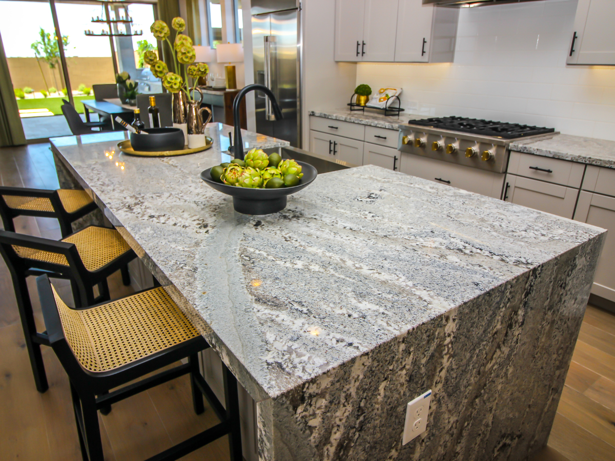 A Countertop Seams Guide to Fabrication and Installation 4 Modern kitchen with a large gray granite island showcasing expertly aligned countertop seams (see our countertop seams guide for tips), black chairs with woven seats, a bowl of green artichokes, and stainless steel appliances in the background.