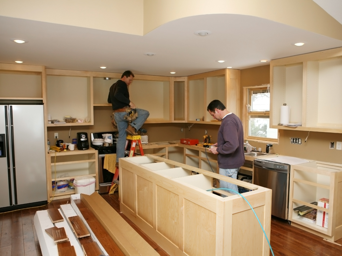 Home Remodel Mistakes to Avoid Before You Start Renovating 3 Two men work on installing cabinets in a kitchen under renovation, carefully addressing remodel mistakes to avoid. Construction tools, a ladder, and unfinished cabinets are visible throughout the space.