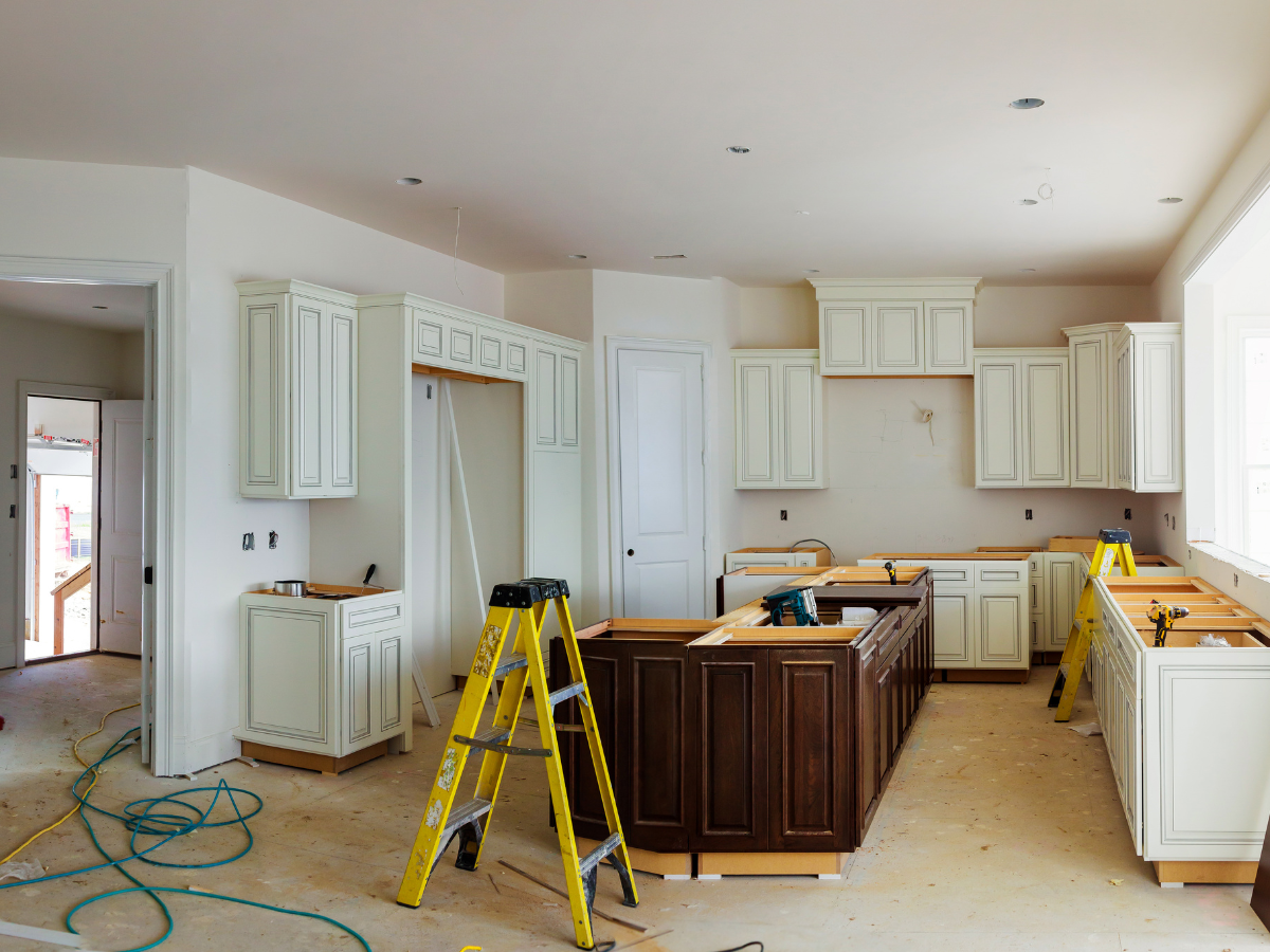 Home Remodel Mistakes to Avoid Before You Start Renovating 5 Kitchen under construction with white cabinets, a dark island, two stepladders, scattered tools, and unfinished flooring—a setting that highlights common remodel mistakes to avoid.