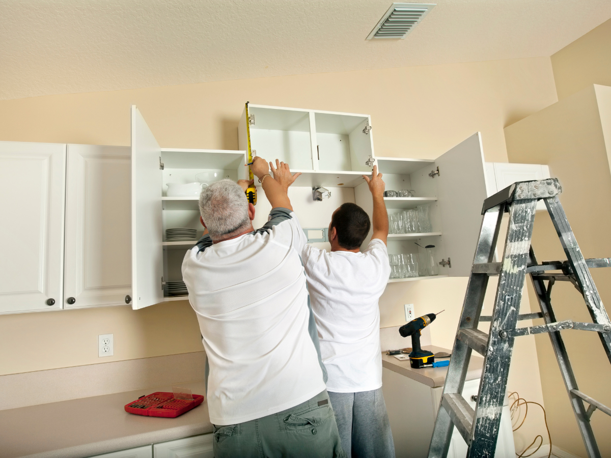 Home Remodel Mistakes to Avoid Before You Start Renovating 6 Two people install a white wall cabinet in a kitchen using a power drill and tools, carefully positioning it to ensure accuracy—a step ladder stands nearby as they work, mindful of remodel mistakes to avoid.