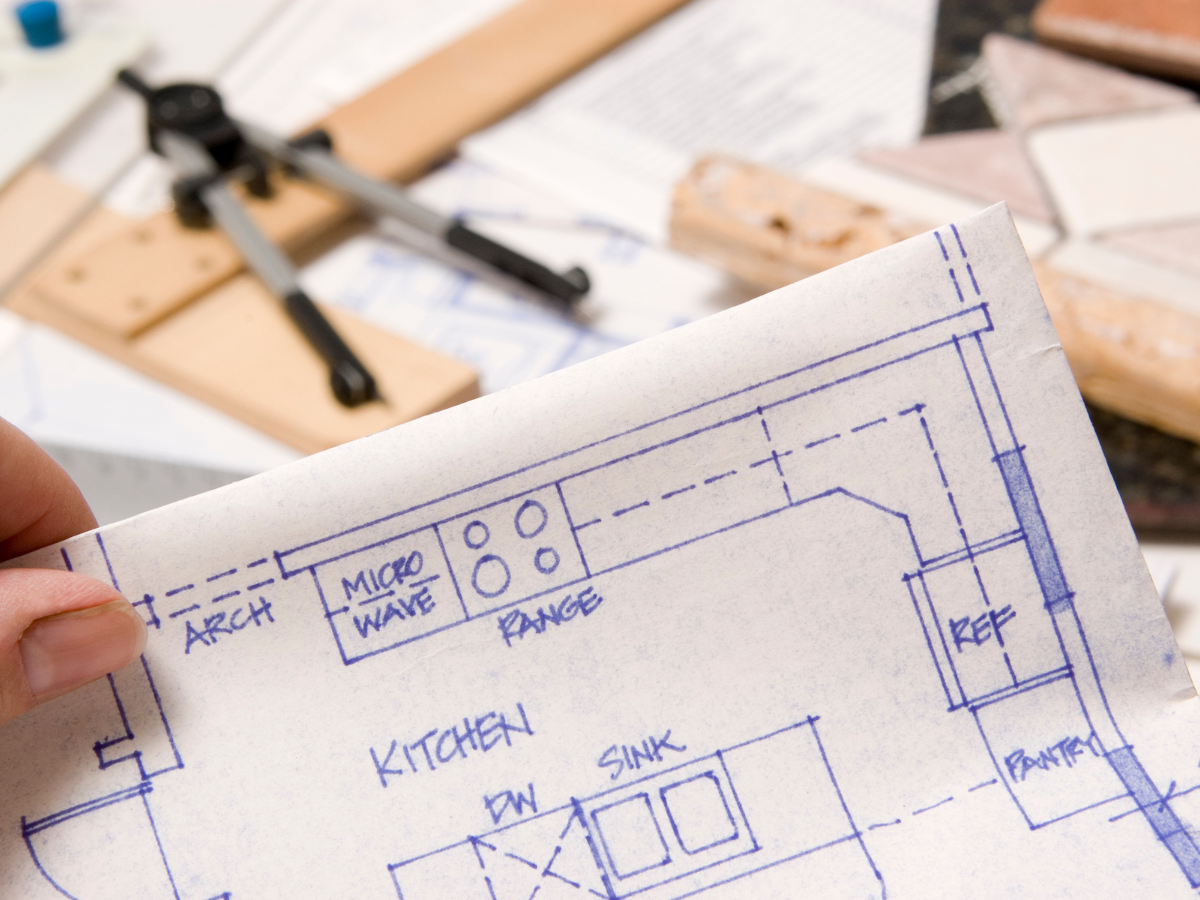 Home Remodel Mistakes to Avoid Before You Start Renovating 1 Hand holding a kitchen blueprint with labeled appliances and counters; compass, tiles, and paperwork are visible in the background, serving as a helpful guide for identifying remodel mistakes to avoid.